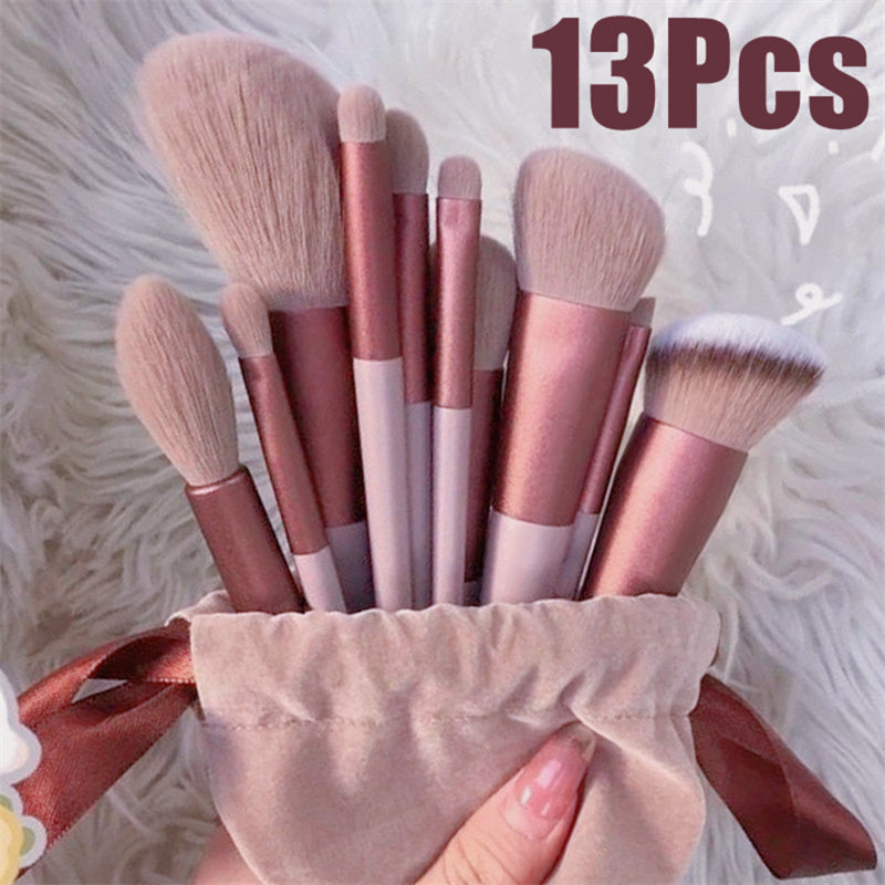 A 13-piece makeup brush set with rose gold and white handles, light pink bristles, and a matching rose gold ribbon, held in a beige velvet pouch against a white faux fur background.