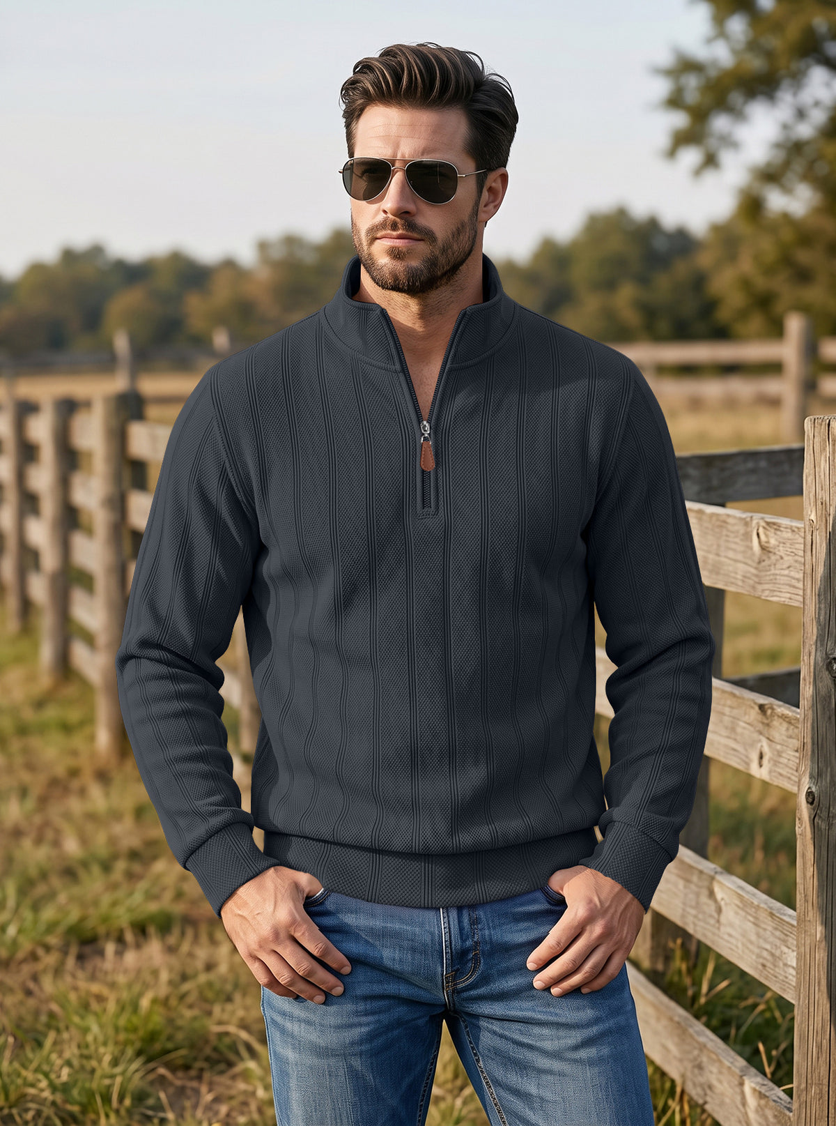 A man wearing a dark charcoal gray quarter-zip pullover sweater and blue jeans, standing outdoors in front of a wooden fence.