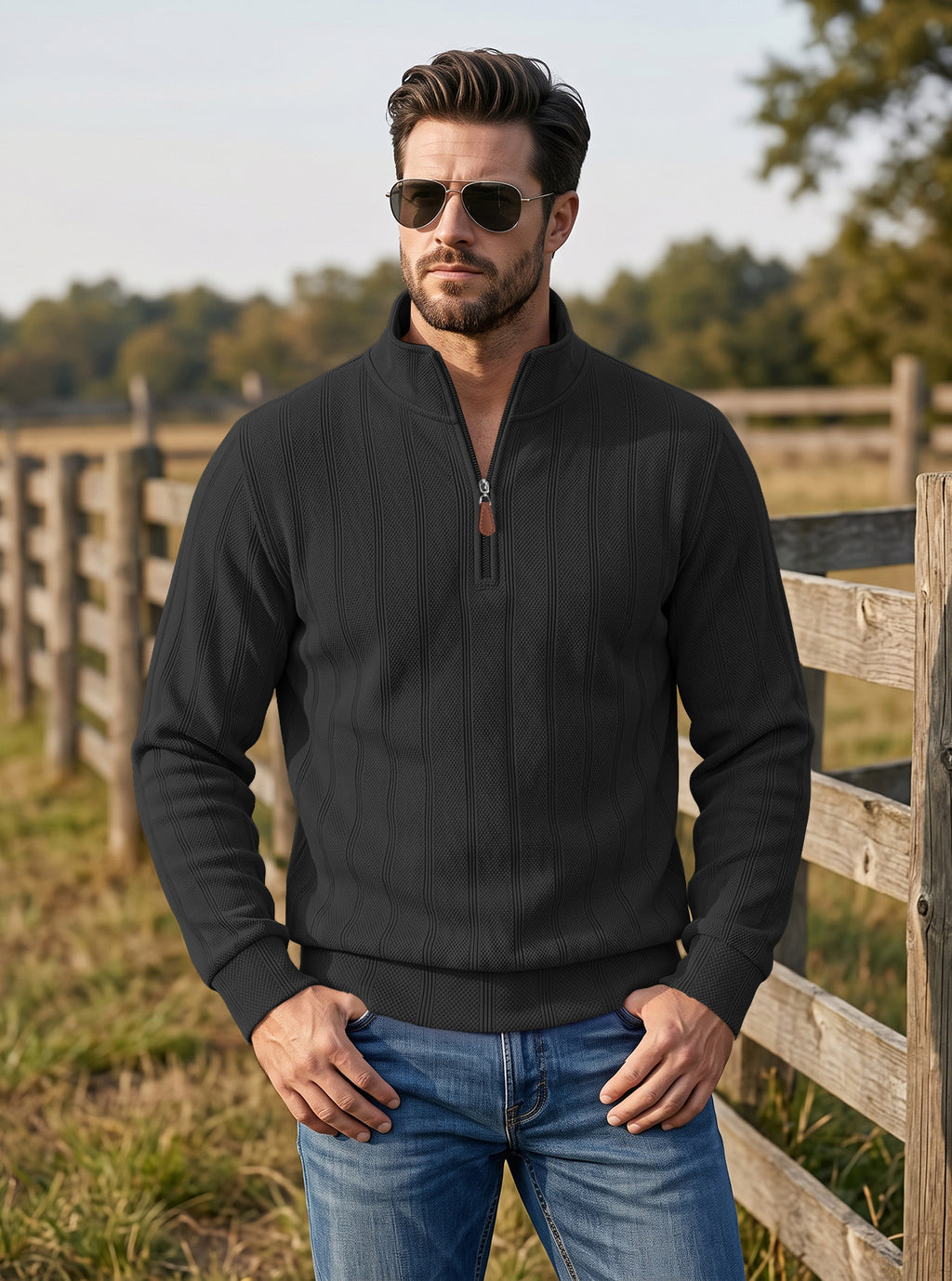 A man wearing a black quarter-zip pullover sweater and blue jeans stands outdoors in front of a wooden fence and trees, wearing dark sunglasses.