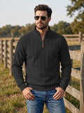 A man wearing a black quarter-zip pullover sweater and blue jeans stands outdoors in front of a wooden fence and trees, wearing dark sunglasses.