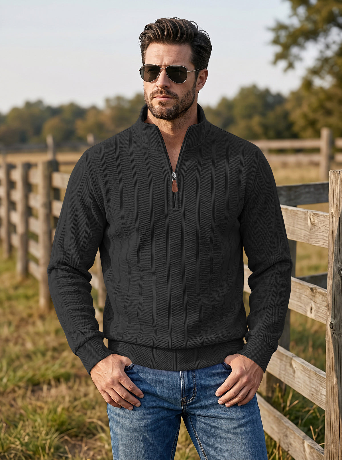 A man wearing a black quarter-zip pullover sweater and blue jeans stands outdoors in front of a wooden fence and trees, wearing dark sunglasses.