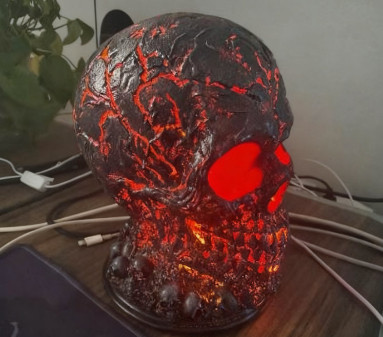 A skull-shaped decorative lamp with a dark textured surface and glowing red internal lights, placed on a wooden surface with cables and a plant in the background.