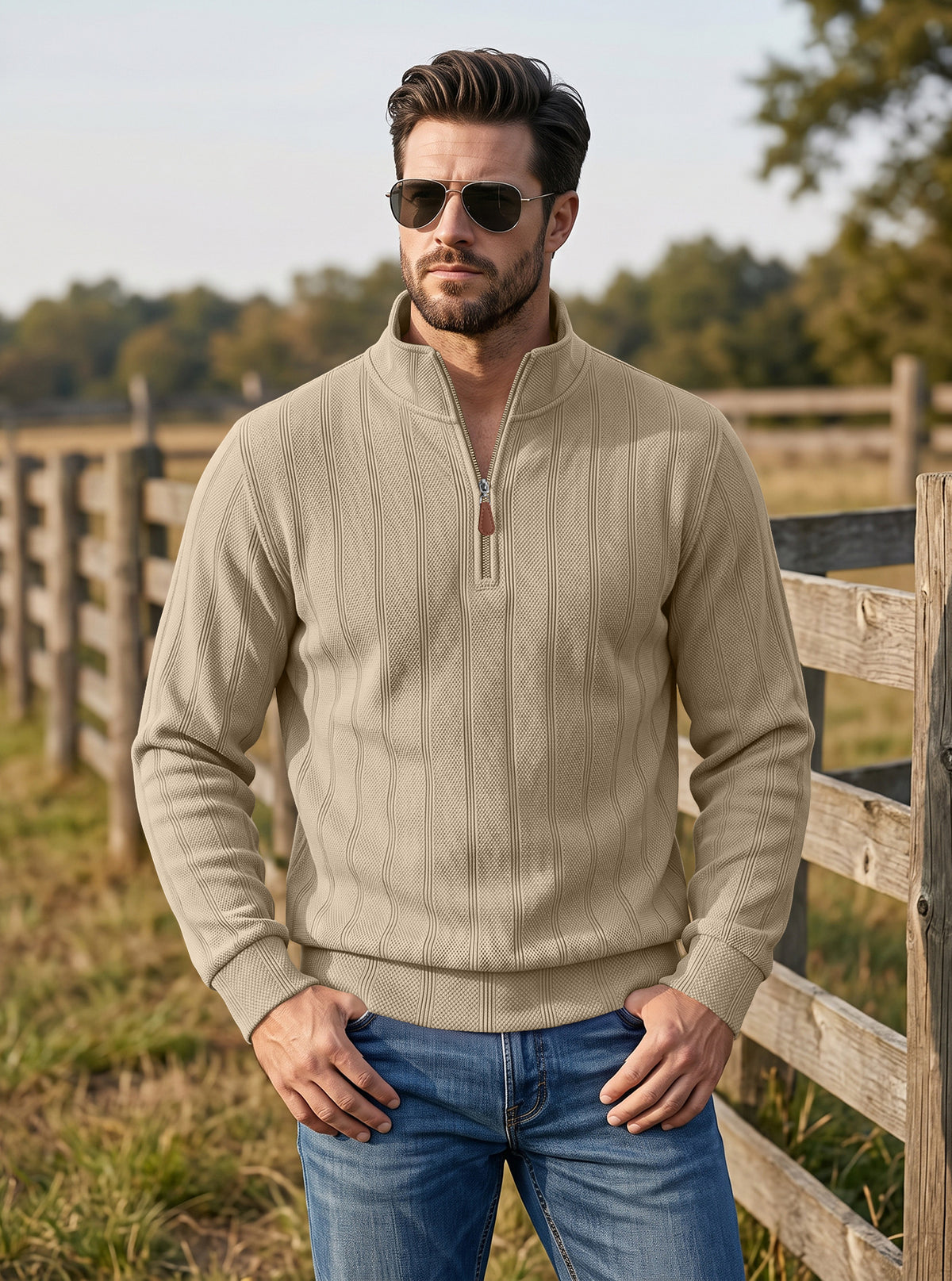 A man wearing a beige ribbed quarter-zip pullover sweater and blue jeans, standing outdoors in front of a wooden fence.