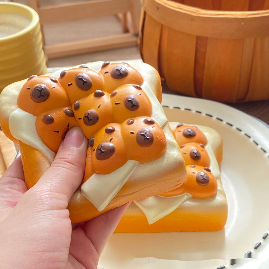 A hand holds a novelty squishy toy shaped like a slice of toast with orange-brown bread, white cream, and small bear faces, with more of the same on a plate in the background.