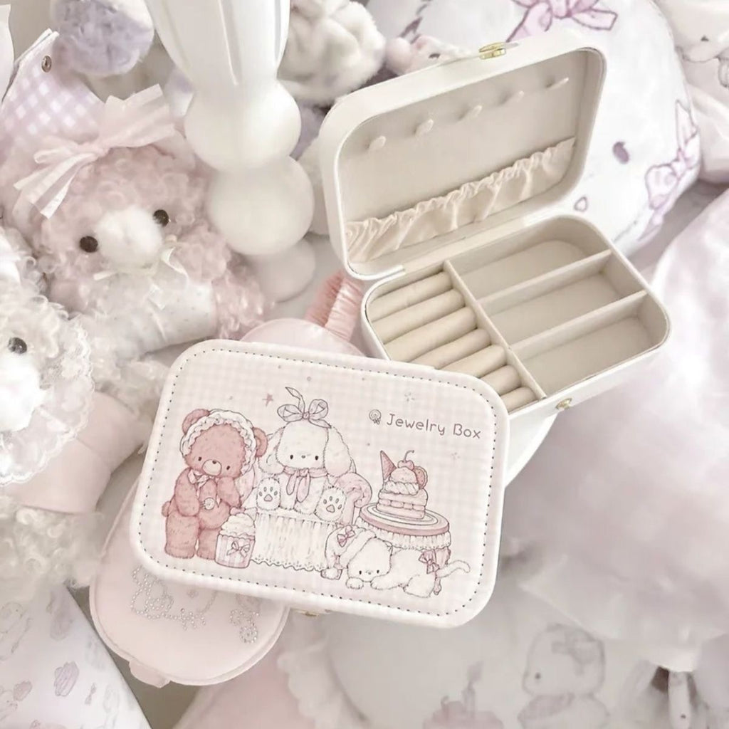 A small cream-colored jewelry box with a cute teddy bear illustration on the front, shown open with interior compartments and rings inside, surrounded by soft plush toys and pastel bedding.