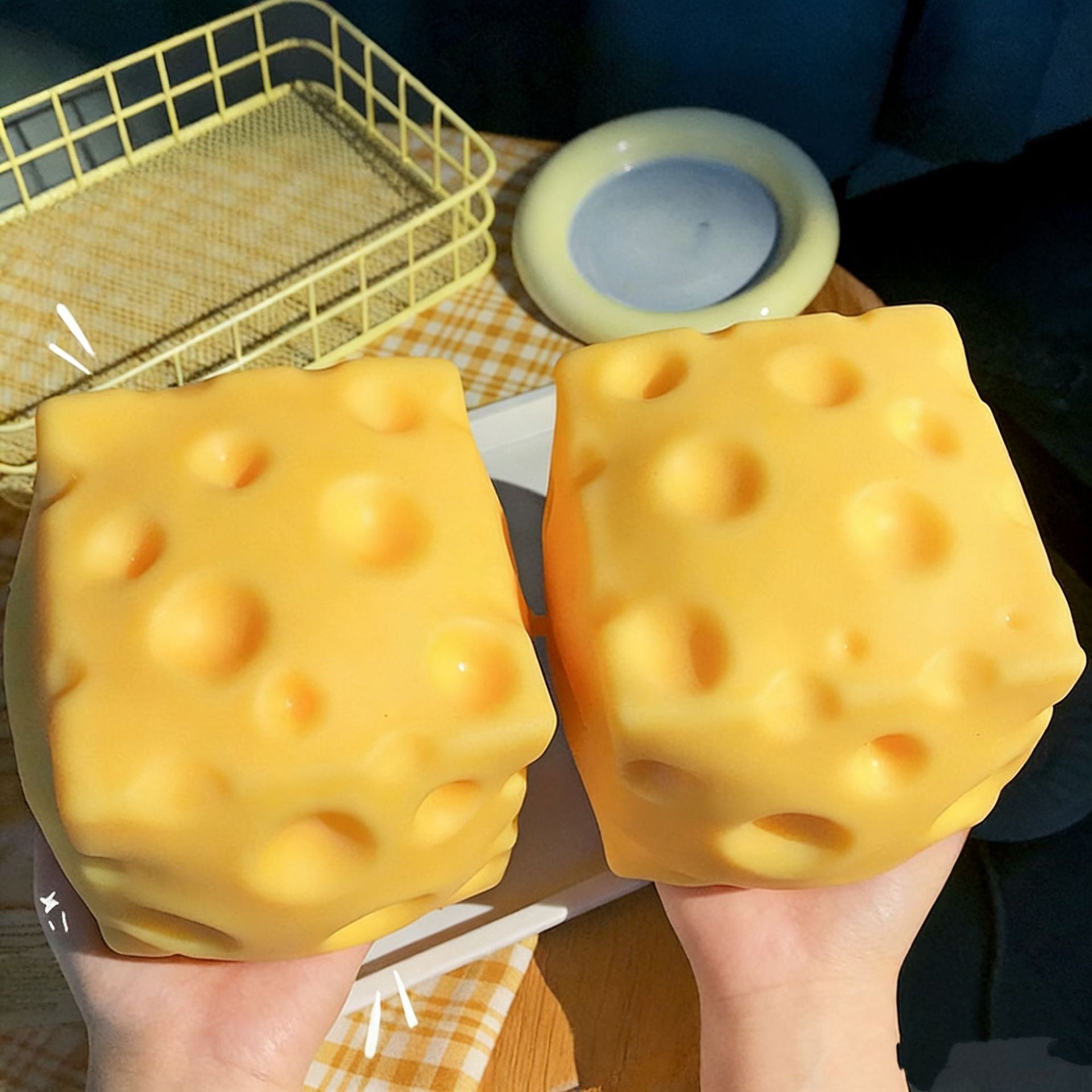 Two large yellow cheese cubes squishy held by hands with a checkered tablecloth and basket in the background.