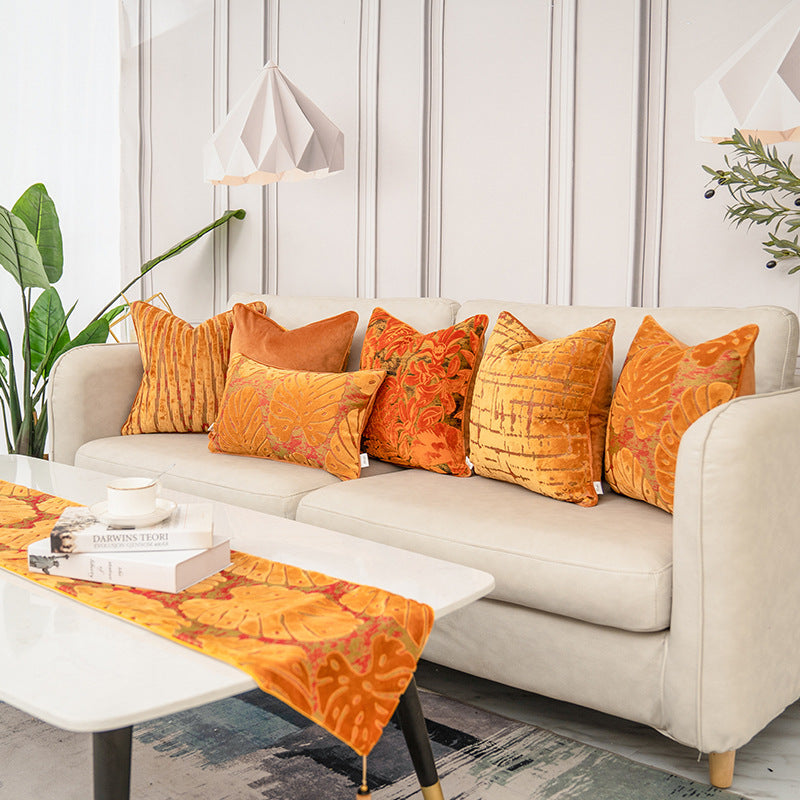 A set of six decorative throw pillows in orange and yellow patterns arranged on a light beige sofa in a modern living room setting.