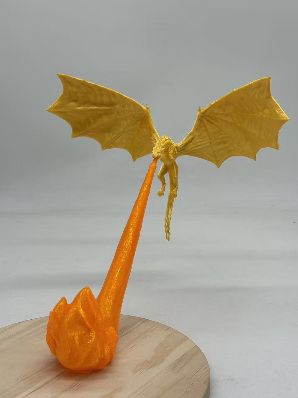 A 3D-printed dragon figurine in yellow with spread wings, perched on a long orange tail-like structure that rests on a round wooden base, photographed against a plain light gray background.