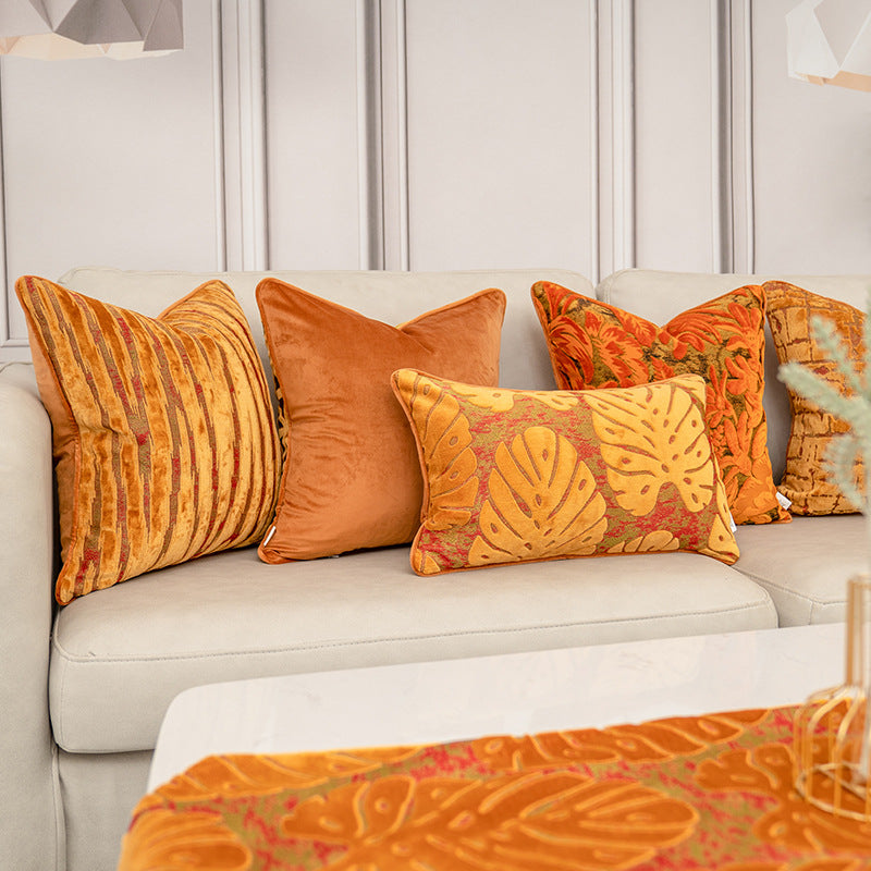 A set of five decorative throw pillows in warm autumn and tropical patterns, styled on a light beige sofa in a modern living room setting.