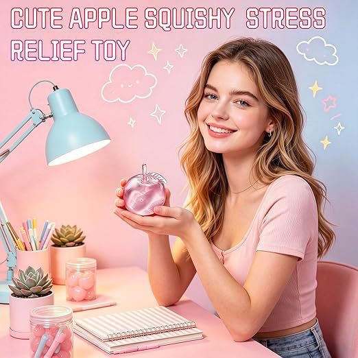 A smiling young woman sits at a pink desk holding a shiny pink apple-shaped stress relief toy. The scene is styled with a light blue desk lamp, notebooks, succulents, and pastel background graphics reading 'CUTE APPLE SQUISHY STRESS RELIEF TOY'.