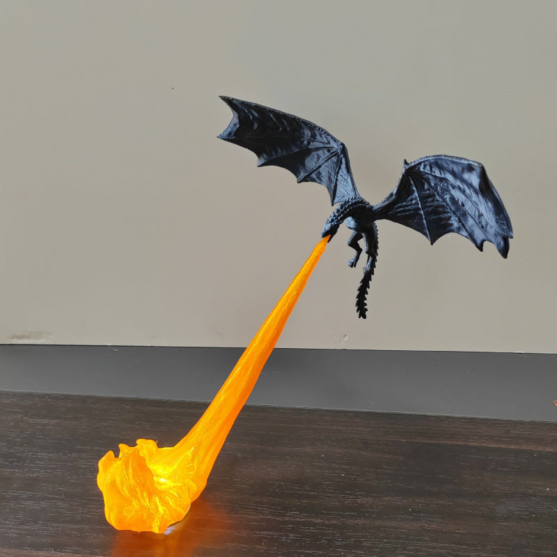 A black dragon figurine with wings spread, breathing a stream of glowing orange flame upward from a base on a dark wooden surface against a plain beige wall.