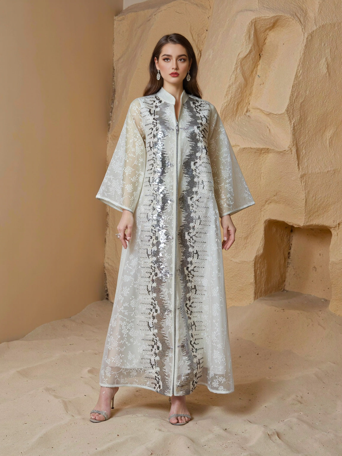 Woman wearing a long, light-colored dress with sequin detailing against a stone wall.