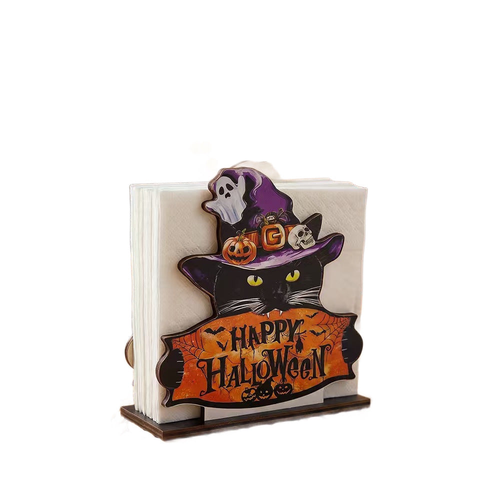A Halloween-themed paper napkin holder featuring a black cat wearing a purple witch hat with a white ghost and orange pumpkins, holding a banner that reads 'Happy Halloween' above a stack of white napkins on a wooden stand.