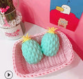 Two green pineapple-shaped squishy toy on a pink woven tray with a pink background and decorative elements.