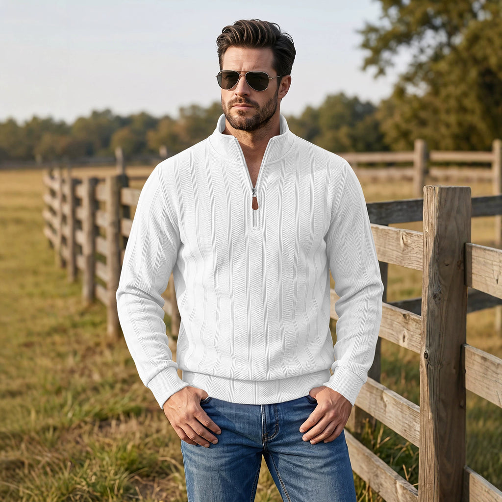 A man wearing a white ribbed quarter-zip pullover sweater and blue jeans stands outdoors in a grassy field beside a wooden fence.