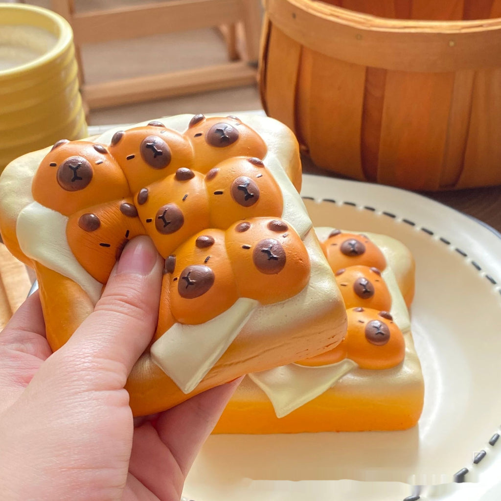 A hand holds a novelty squishy toy shaped like a slice of toast with orange-brown bread, white cream, and small bear faces, with more of the same on a plate in the background.
