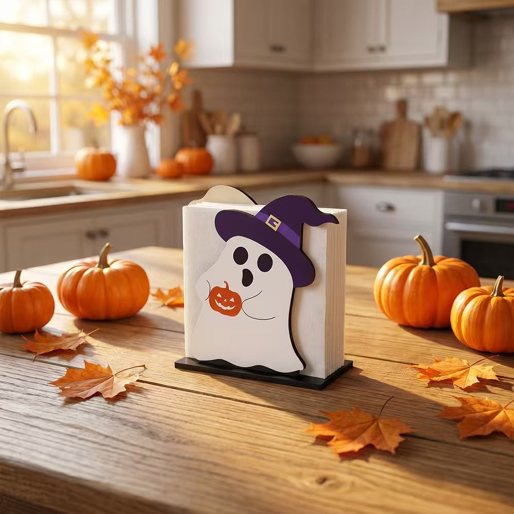 A Halloween-themed wooden napkin holder featuring a white ghost with a purple witch hat and holding a jack-o'-lantern, standing on a wooden kitchen table surrounded by pumpkins and autumn leaves.