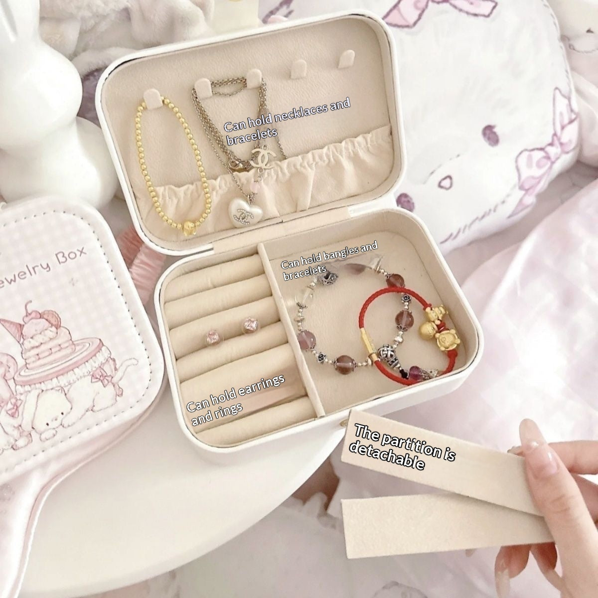 An open round jewelry box with cream-colored interior compartments holding bracelets, rings, and earrings, shown on a soft pink surface. A hand holds a small card next to the box.