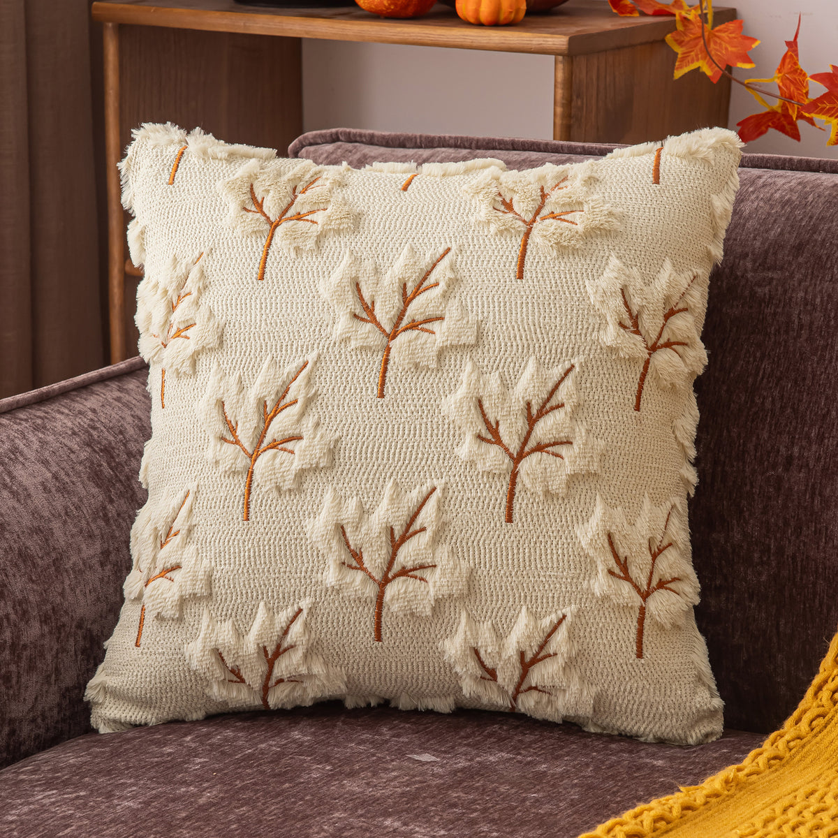 A square cream throw pillow with raised autumn leaf embroidery, resting on a dark brown upholstered sofa in a cozy indoor setting.