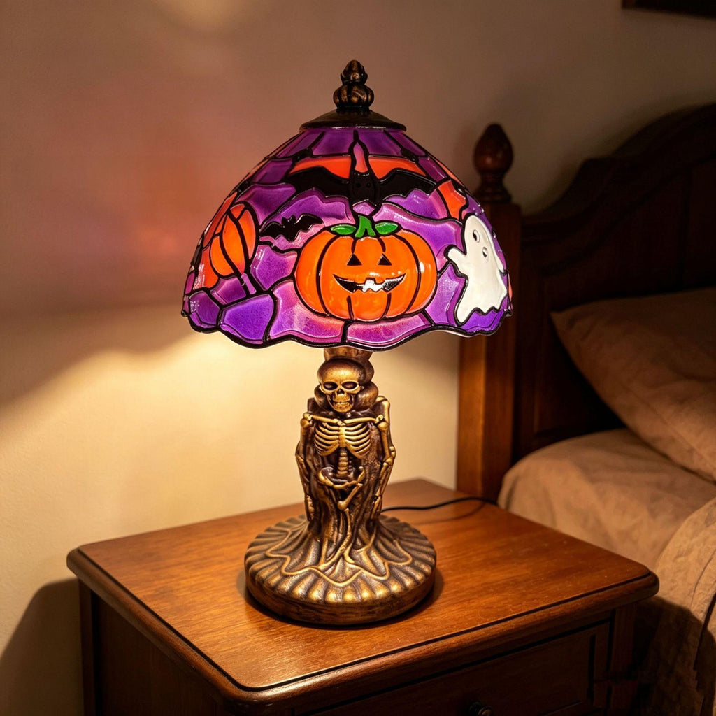 A Halloween-themed table lamp with a stained-glass shade featuring a jack-o'-lantern, bats, and a ghost, set on a wooden nightstand in a bedroom.