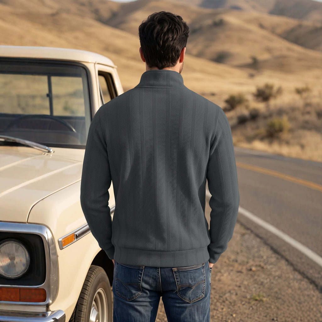 A man viewed from behind wearing a dark gray ribbed quarter-zip pullover sweater and blue jeans, standing next to a vintage cream pickup truck on a desert road with rolling hills in the background.