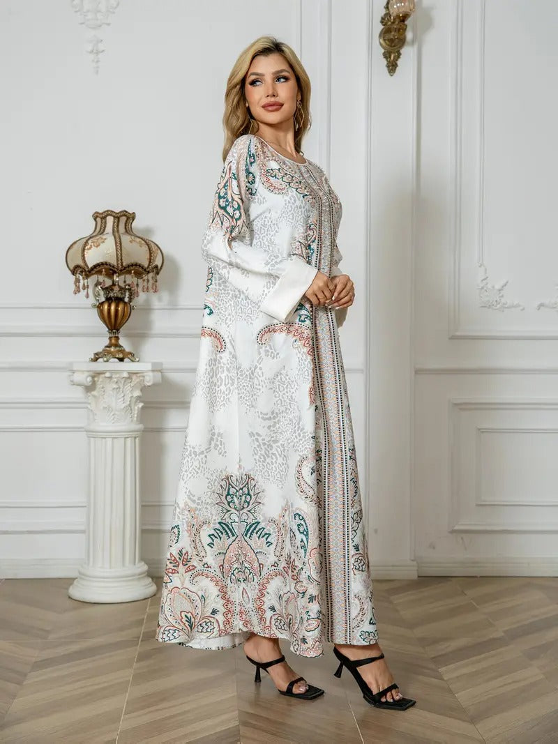 A woman models a long white maxi dress with intricate multicolor paisley and floral patterns in teal, pink, and beige, standing in a classic white-paneled room with a wooden floor.