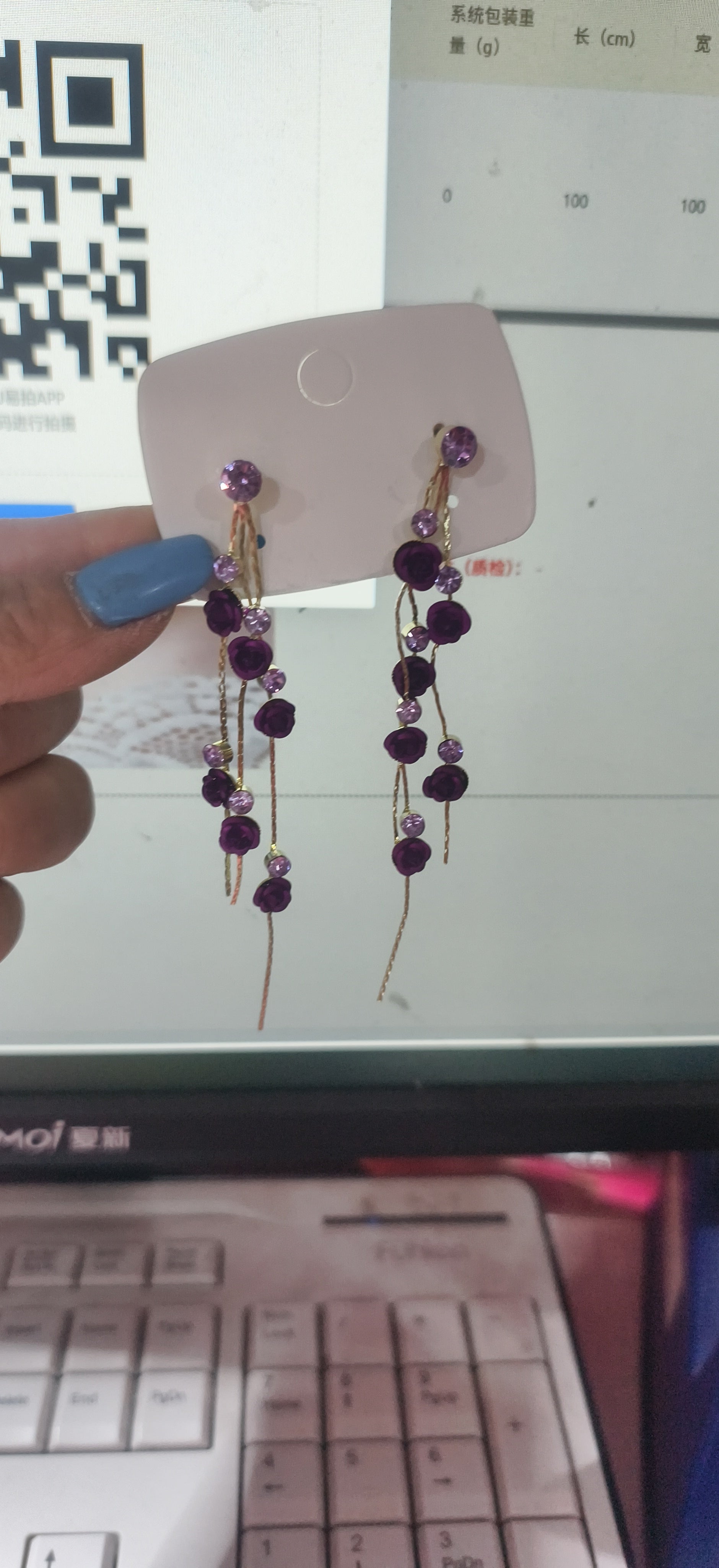A pair of dangling earrings with purple faceted beads and gold-toned chains, displayed on a small pink card held by a hand with blue nail polish in front of a computer monitor and keyboard.