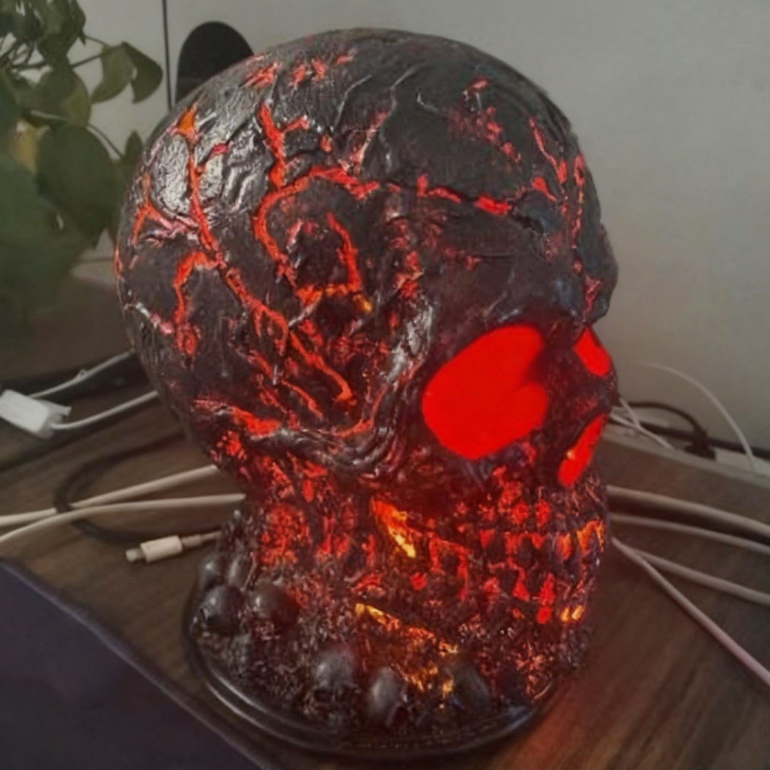 A skull-shaped decorative lamp with a textured black and dark gray surface, glowing red eye sockets, and warm orange-red internal lighting, shown on a desk with cables in the background.