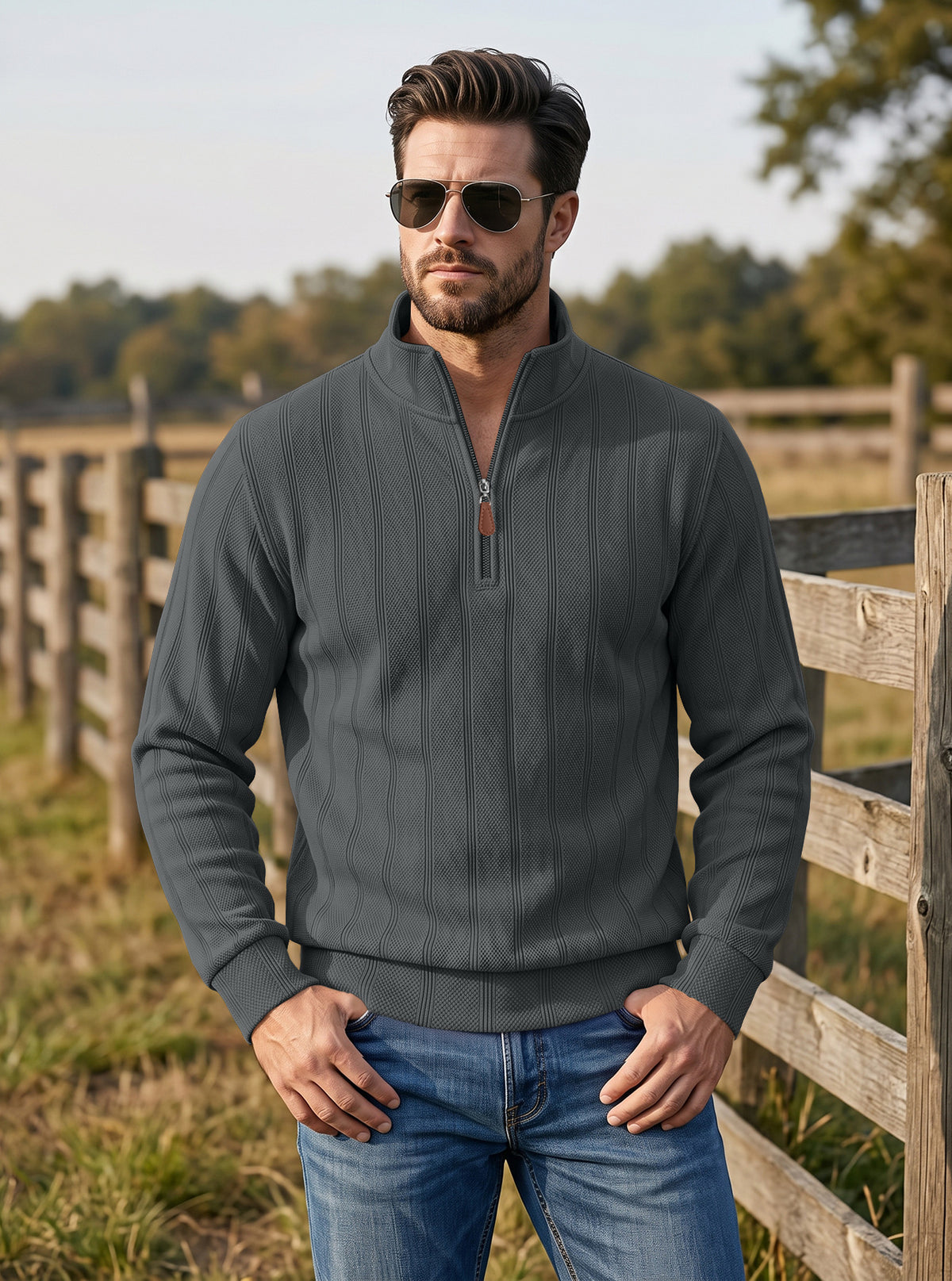 A man wearing a dark gray ribbed quarter-zip pullover sweater and blue jeans, standing outdoors in front of a wooden fence.