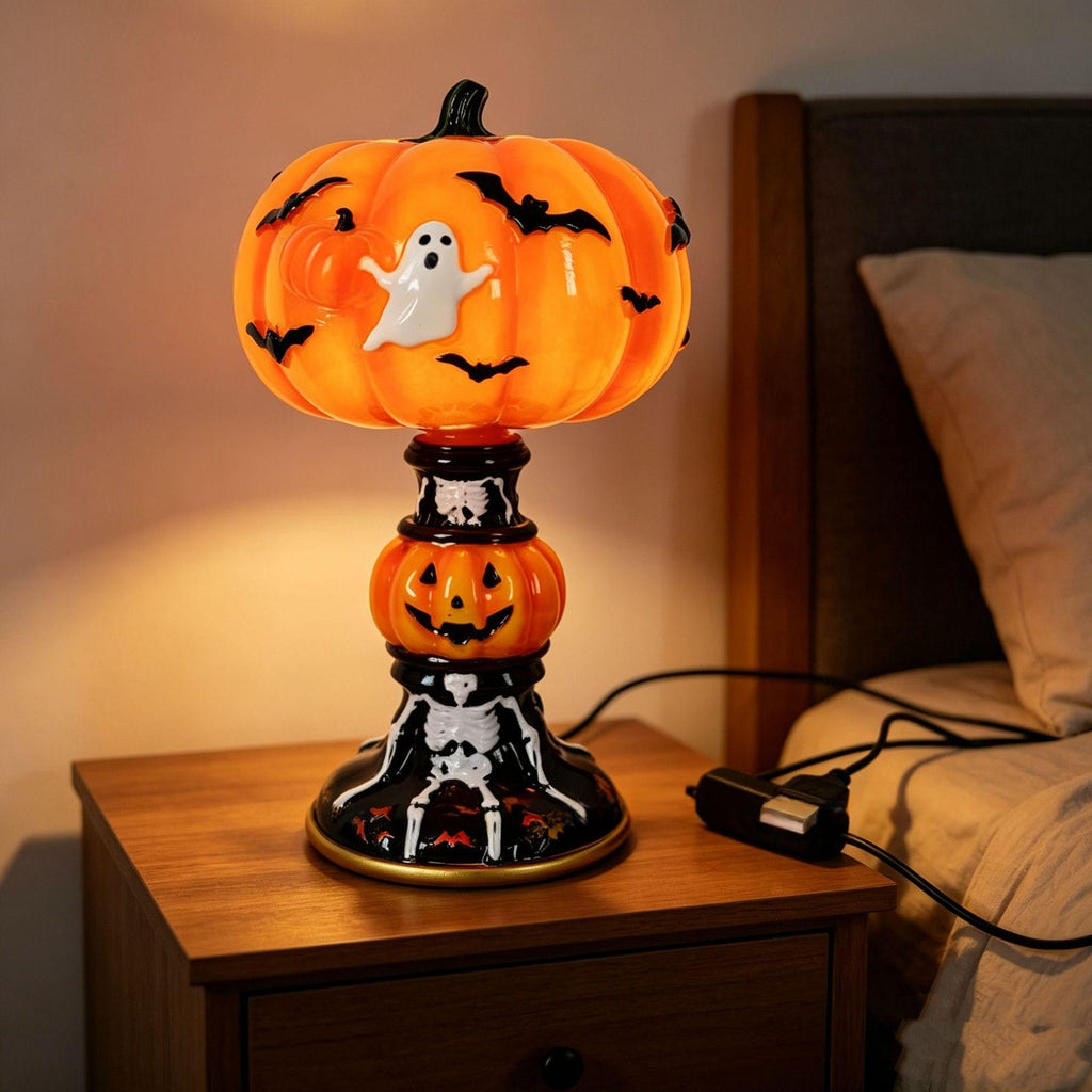 A Halloween-themed table lamp shaped like a glowing orange pumpkin with a white ghost and black bats, set on a wooden bedside table next to a bed.