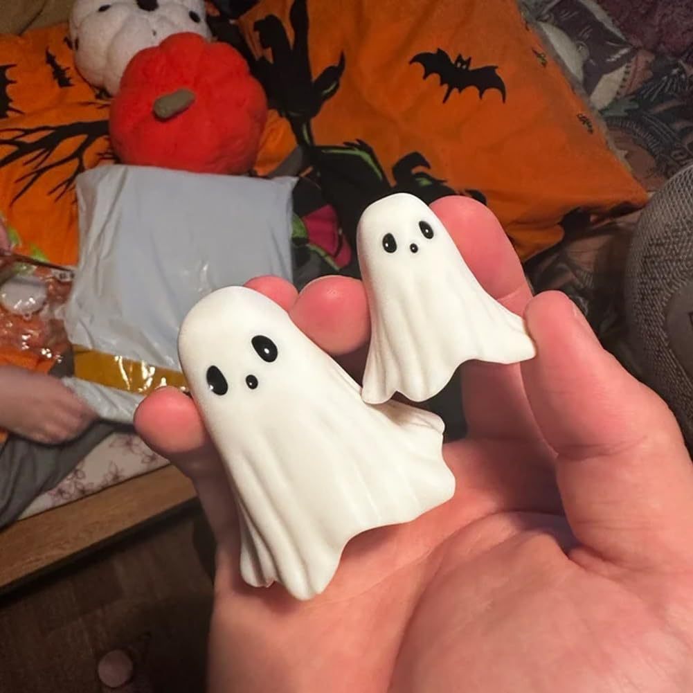 Two small ghost figurines held in a hand with Halloween decorations in the background.