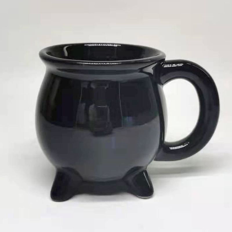 A glossy black ceramic mug shaped like a small cauldron with three feet and a large handle, shown on a plain light background.