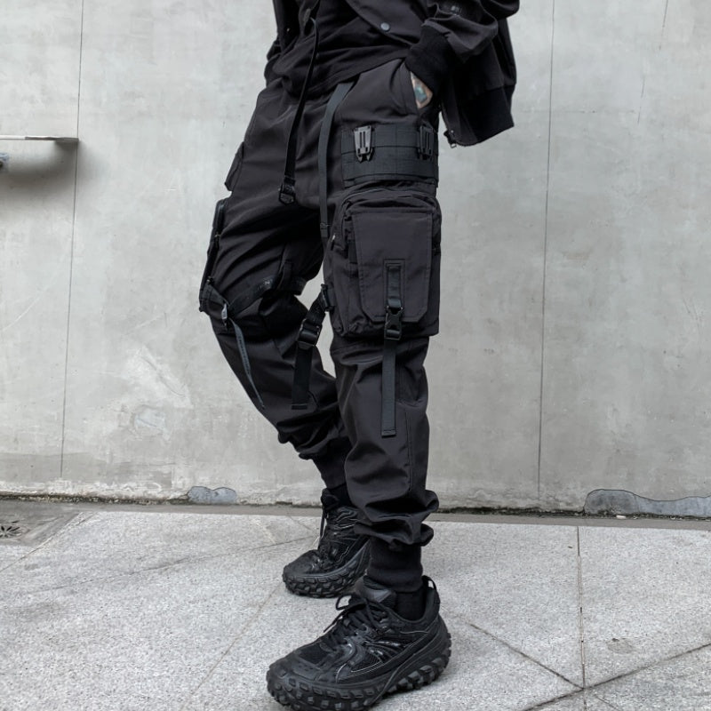 Person wearing black tactical pants and shoes against a gray wall.