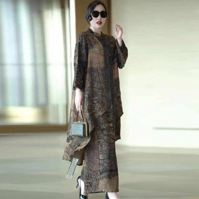 A woman models a long-sleeved, loose-fitting maxi dress with an abstract, painterly pattern in earthy brown, beige, and black tones. She is wearing black sunglasses, holding a small blue-green handbag, and standing in a brightly lit indoor space with a soft-focus background.