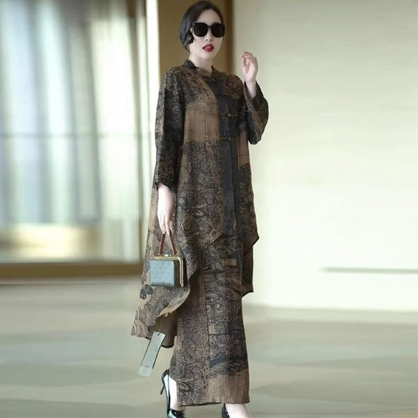 A woman models a long-sleeved, loose-fitting maxi dress with an abstract, painterly pattern in earthy brown, beige, and black tones. She is wearing black sunglasses, holding a small blue-green handbag, and standing in a brightly lit indoor space with a soft-focus background.