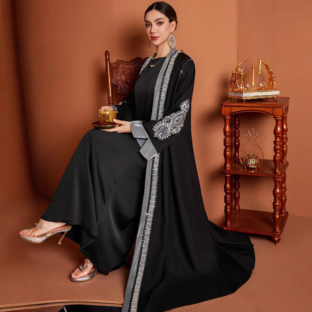 A woman seated in a warm brown studio setting wearing a black abaya with a long sheer gray sleeve and ornate silver embroidery, holding a small tray. A wooden chair and matching side table are visible behind her.