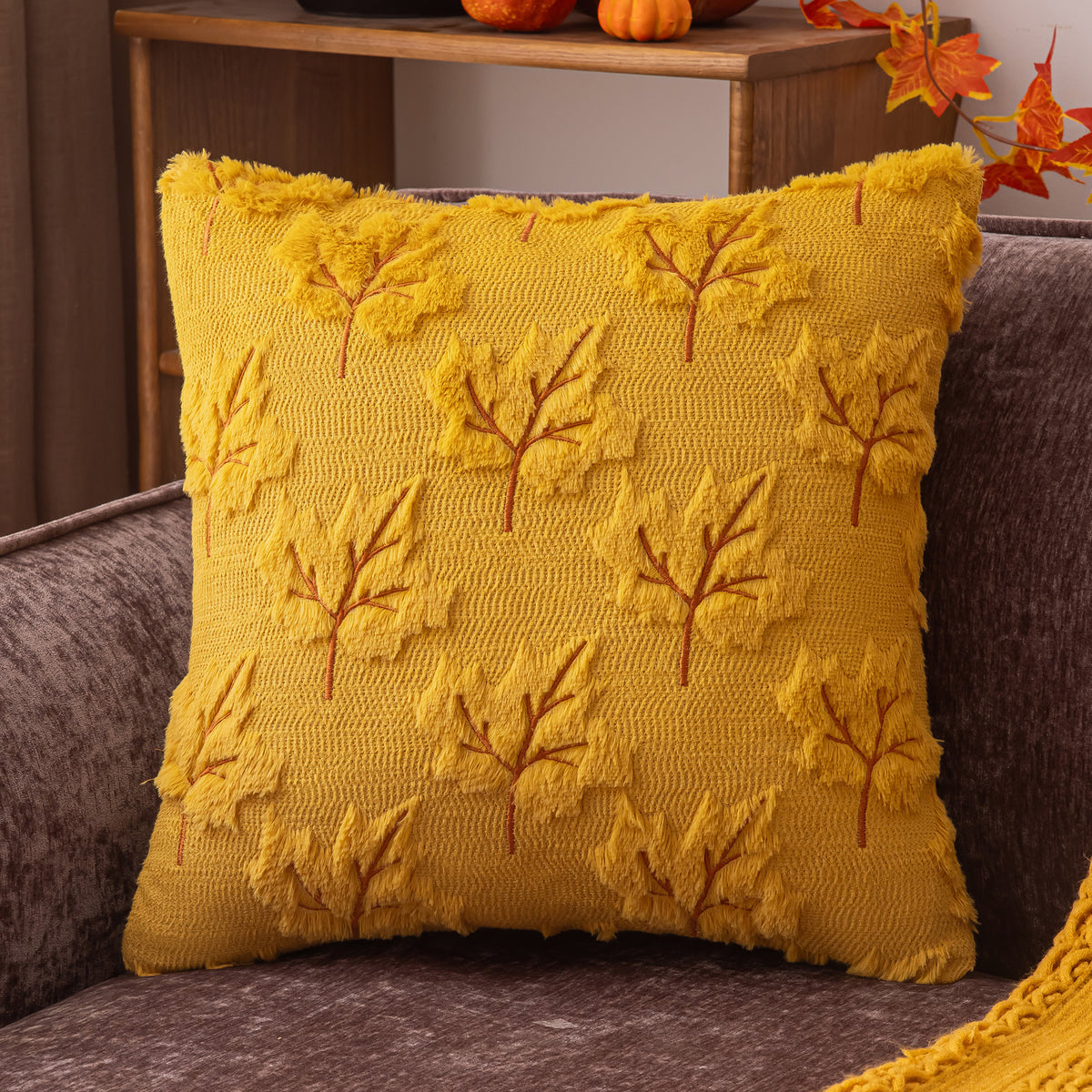 A square yellow throw pillow with a textured knit leaf pattern, resting on a gray upholstered chair in a cozy indoor setting.