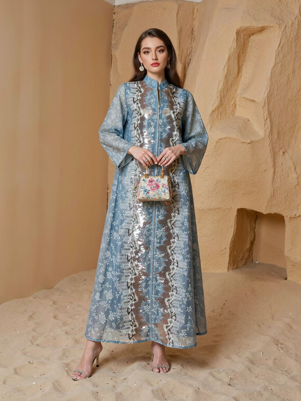 Woman in a long, light blue embroidered dress standing in a sandy area with beige rock formations.