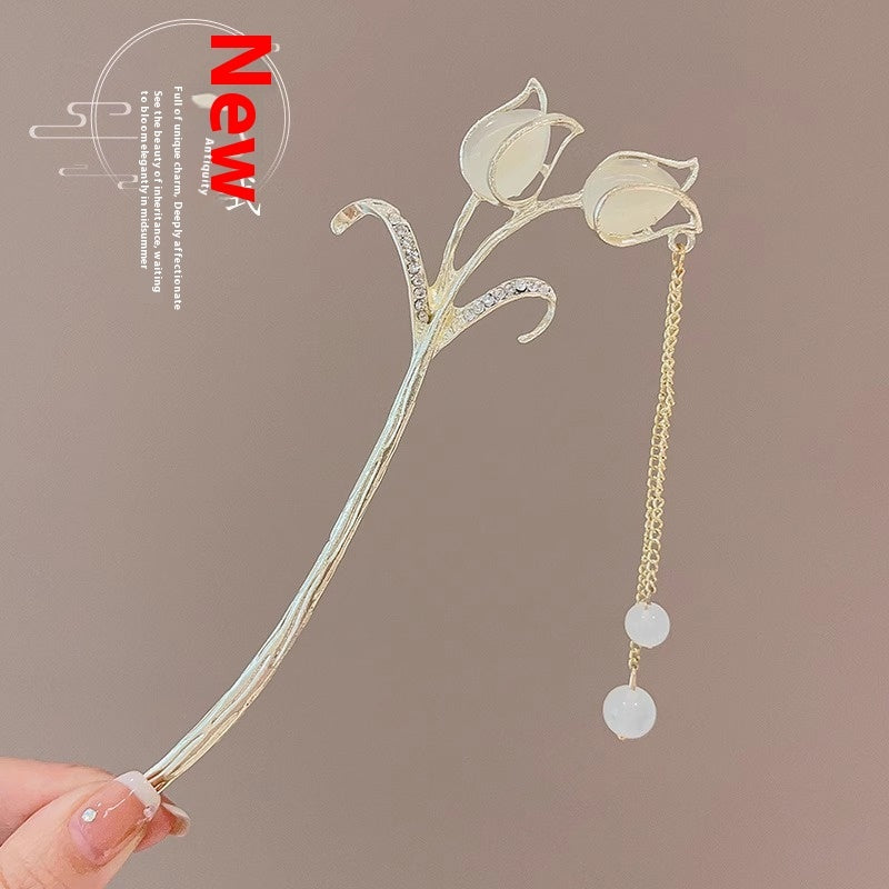 Decorative hairpin with floral design and chain tassel held by a hand against a brown background.