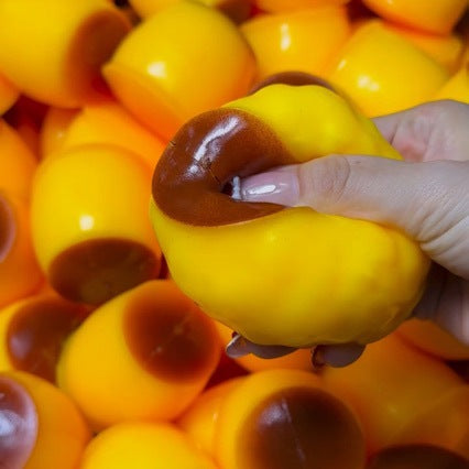 Hand holding a yellow pudding squishy with a brown stem against a background of similar fruits.