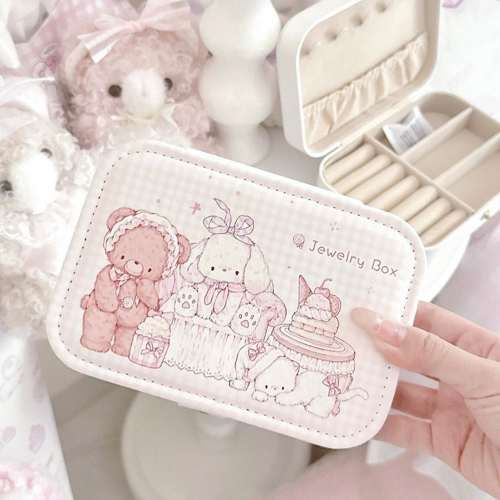 A hand holds a small rectangular jewelry box with rounded corners. The box is white with a soft pink gingham border and a cute illustration of teddy bears, a bunny, and a cake, with the words 'Jewelry Box' printed on the front.