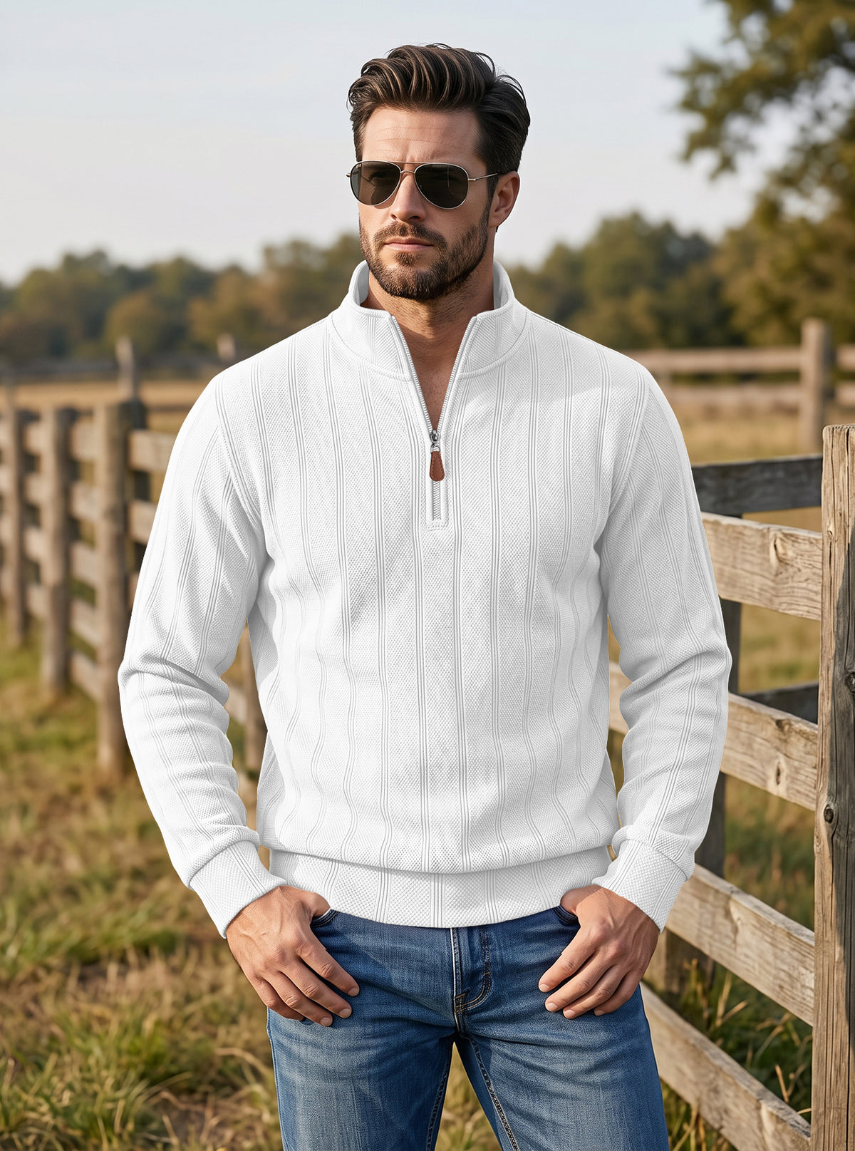 A man wearing a white quarter-zip pullover sweater and blue jeans stands outdoors in front of a wooden fence and trees, wearing dark sunglasses.