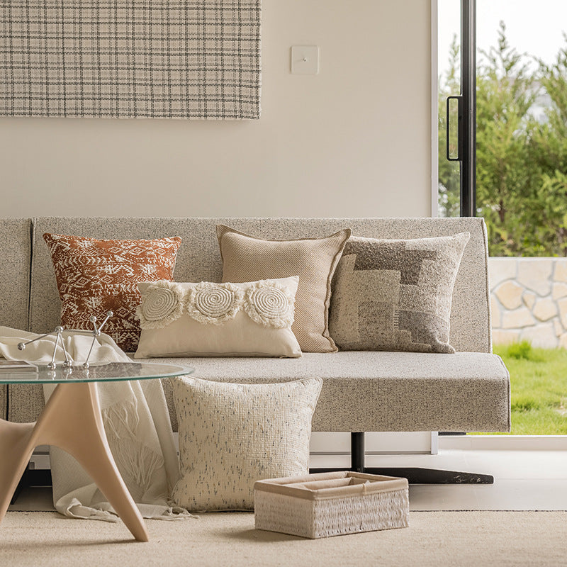 A modern light gray fabric sofa styled with four decorative throw pillows in neutral and warm tones, set in a bright living room with a glass coffee table and a large window showing greenery outside.
