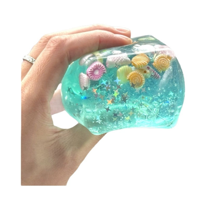 Hand holding a clear container filled with blue slime, colorful sea creature toys, and decorative elements on a white background.