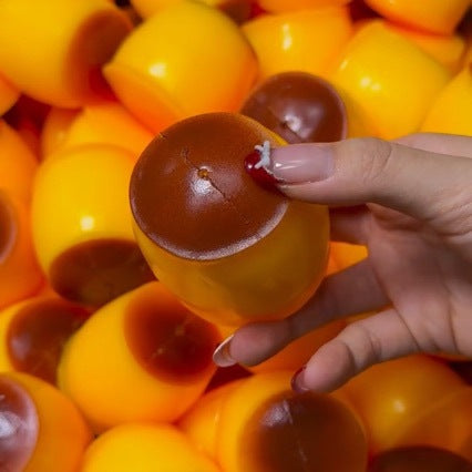 A hand holds a small, round, yellow and brown acorn-shaped novelty toy or capsule, surrounded by a pile of identical yellow and brown capsules.