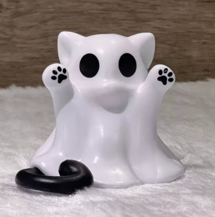 A white ghost-shaped cat figurine with black paw prints on its paws and a black curled tail, standing on a white textured surface against a brown wooden background.