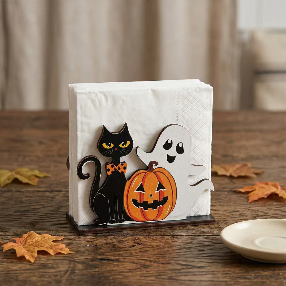 A Halloween-themed paper napkin holder on a rustic wooden table, featuring a black cat with yellow eyes, a white ghost, and an orange jack-o'-lantern pumpkin.