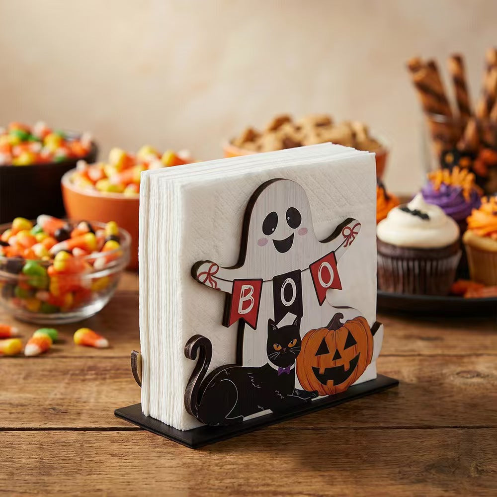 Halloween-themed paper napkins on a black stand, featuring a smiling ghost holding a 'BOO' banner, a black cat, and a jack-o'-lantern, set on a wooden table with candy and cupcakes blurred in the background.