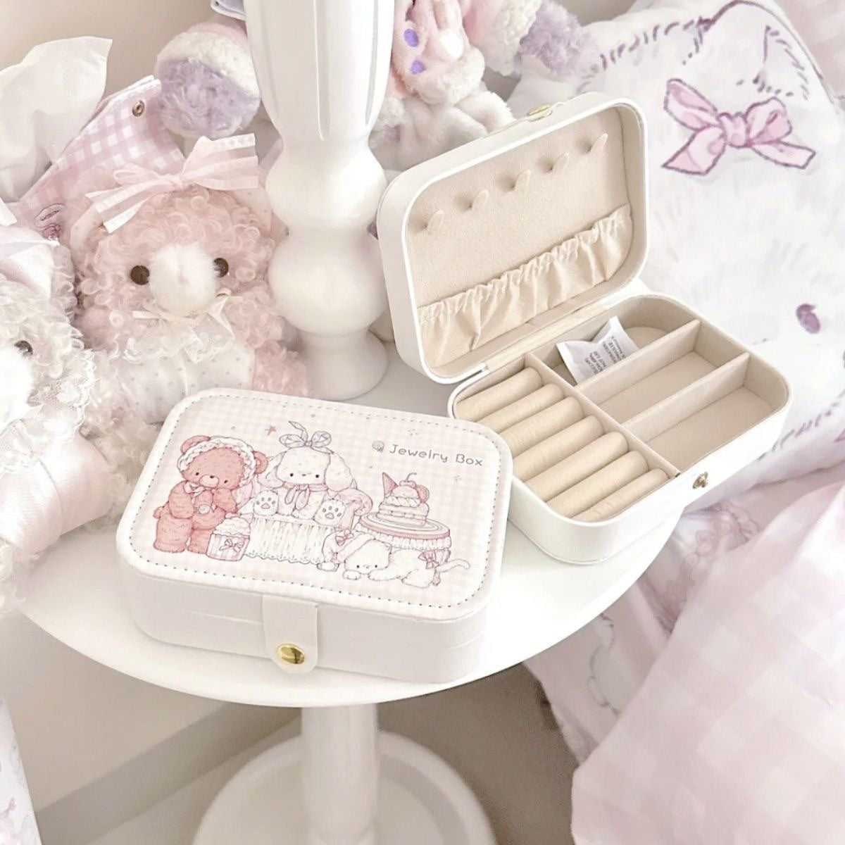 A small cream-colored jewelry box with a cute teddy bear illustration on the front, shown closed on a white table. Another open view of the same box is shown behind it, displaying interior compartments and ring rolls.