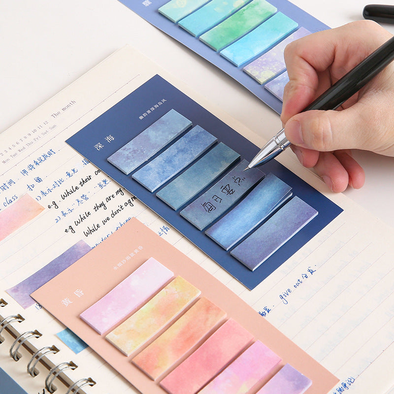 Colorful sticky notes with watercolor design on a notebook page, being used by a hand with a pen.